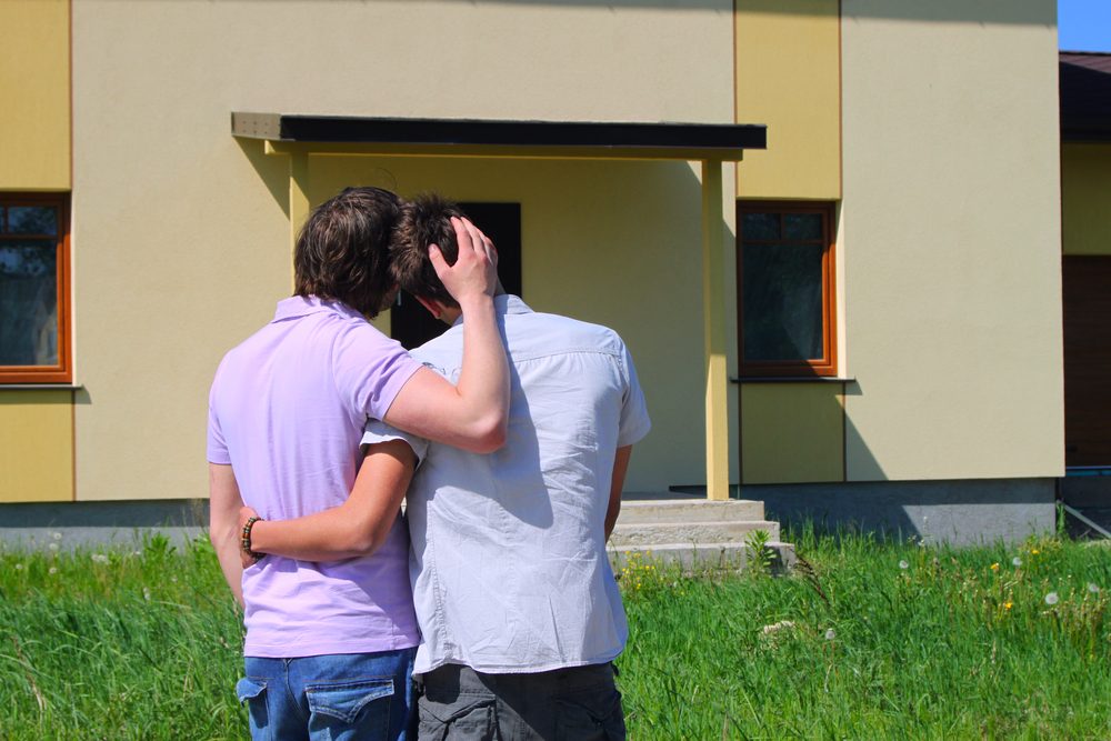 A homosexual couple embrace one another in front of their home. 